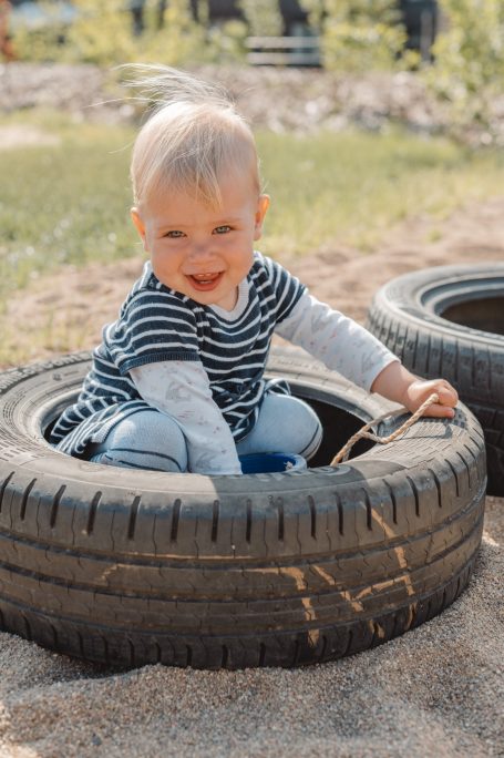 Ein Kleinkind sitzt lächelnd in einem alten Reifen auf einem Spielplatz.