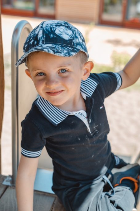 Ein lächelnder Junge in einem gestreiften Shirt und einer Kappe sitzt auf einem Spielplatz.