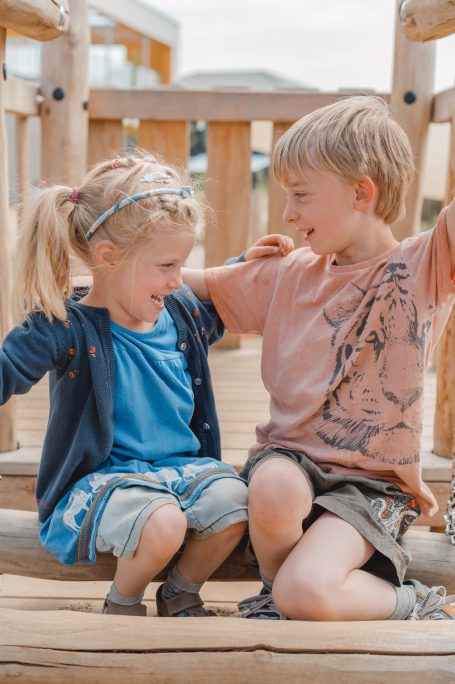 Zwei Kinder lachen und sitzen auf einer Holzspielplatzstruktur.