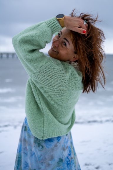 Frau mit lockigem, braunem Haar in grünem Pullover am Strand, lächelt über die Schulter.