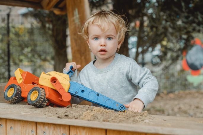 Kleines Kind spielt mit einem bunten Spielzeug-Traktor im Sand.