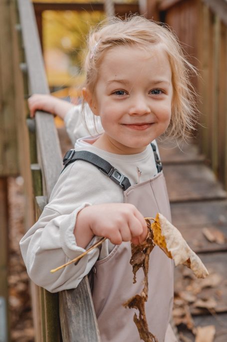 Mädchen mit blonden Haaren hält Laub in der Hand und lächelt auf einer Holzbrücke.